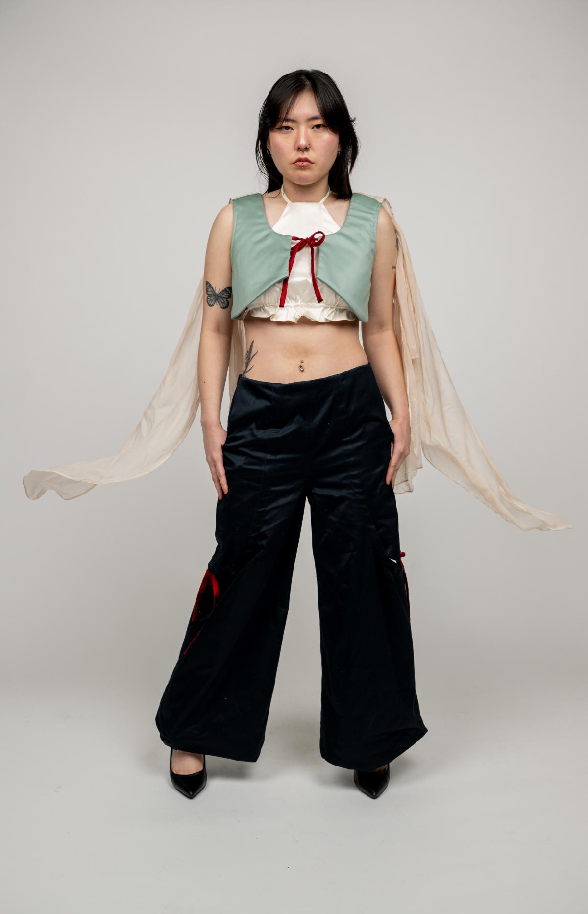 a model wears a cream halter top with a ruffle hem underneath a green cropped vest with a sheer cape. paired with black wide-legged pants