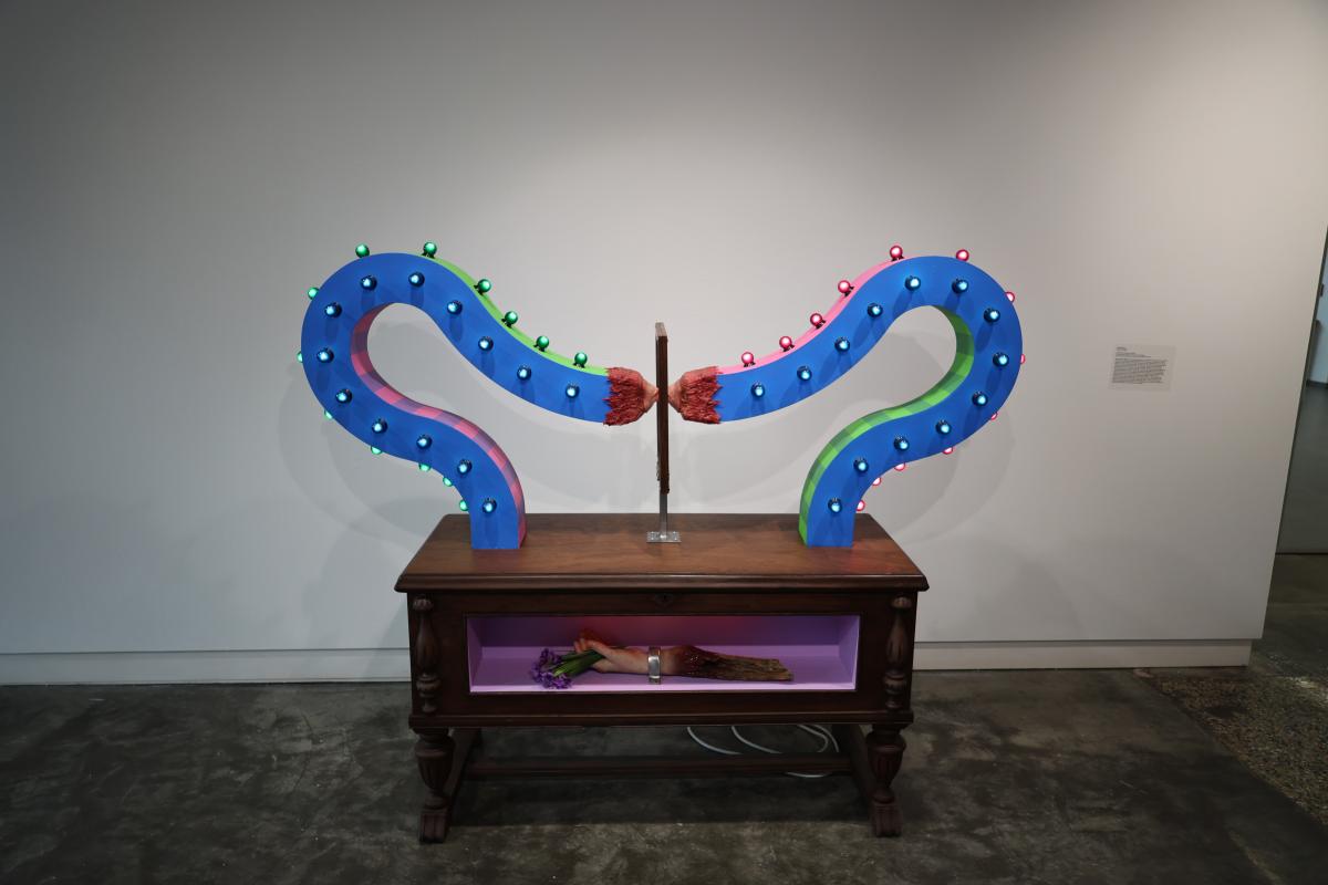 Mixed media piece featuring two mouthes kissing from extended glass backs over a wooden table