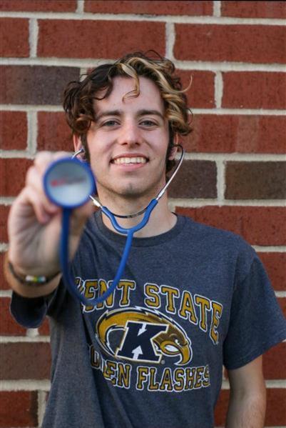 Honors College student Austin Rykowski poses in front of a  brick building in a Kent State shirt holding a stethoscope. PHOTO COURTESY OF .