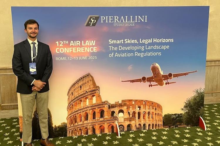 A young man in a blue suit stands in front of a large fabric sign announcing a law conference in Rome, Italy.