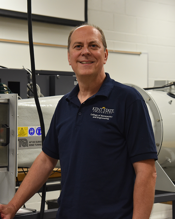 A middle-aged man wearing a blue t-shirt and standing next to an experimental wind tunnel.