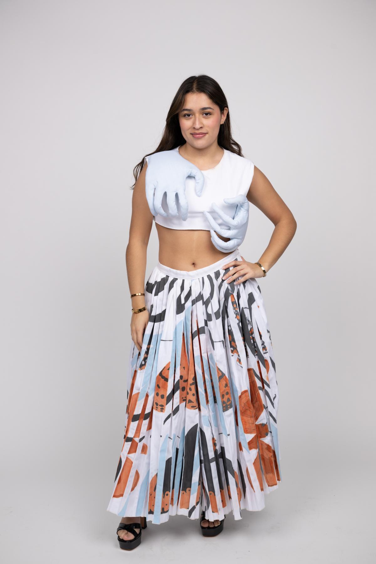 the model wears a white crop top with 3d hands on either side of the garment with a floor length pleated skirt with abstract blue and orange shapes