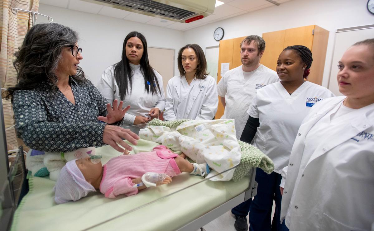 Instructor demonstrates infant care using a lifelike baby simulator while a group of nursing students in clinical uniforms gathers around a hospital bassinet, observing and listening during a hands-on training session in a clinical simulation lab.