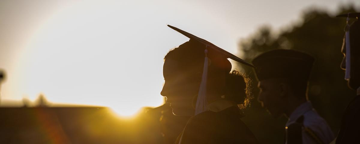 A picture of a young woman in a graduate cap with the sun setting behind her