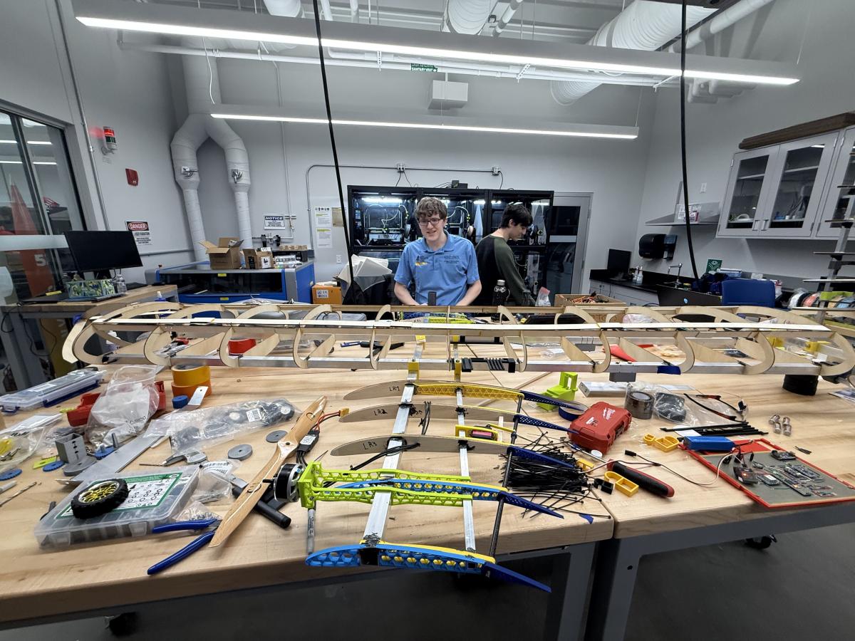 Two male students from Kent State's Design, Build, Fly team stand behind a table in the lab full of all the aircraft components during production.