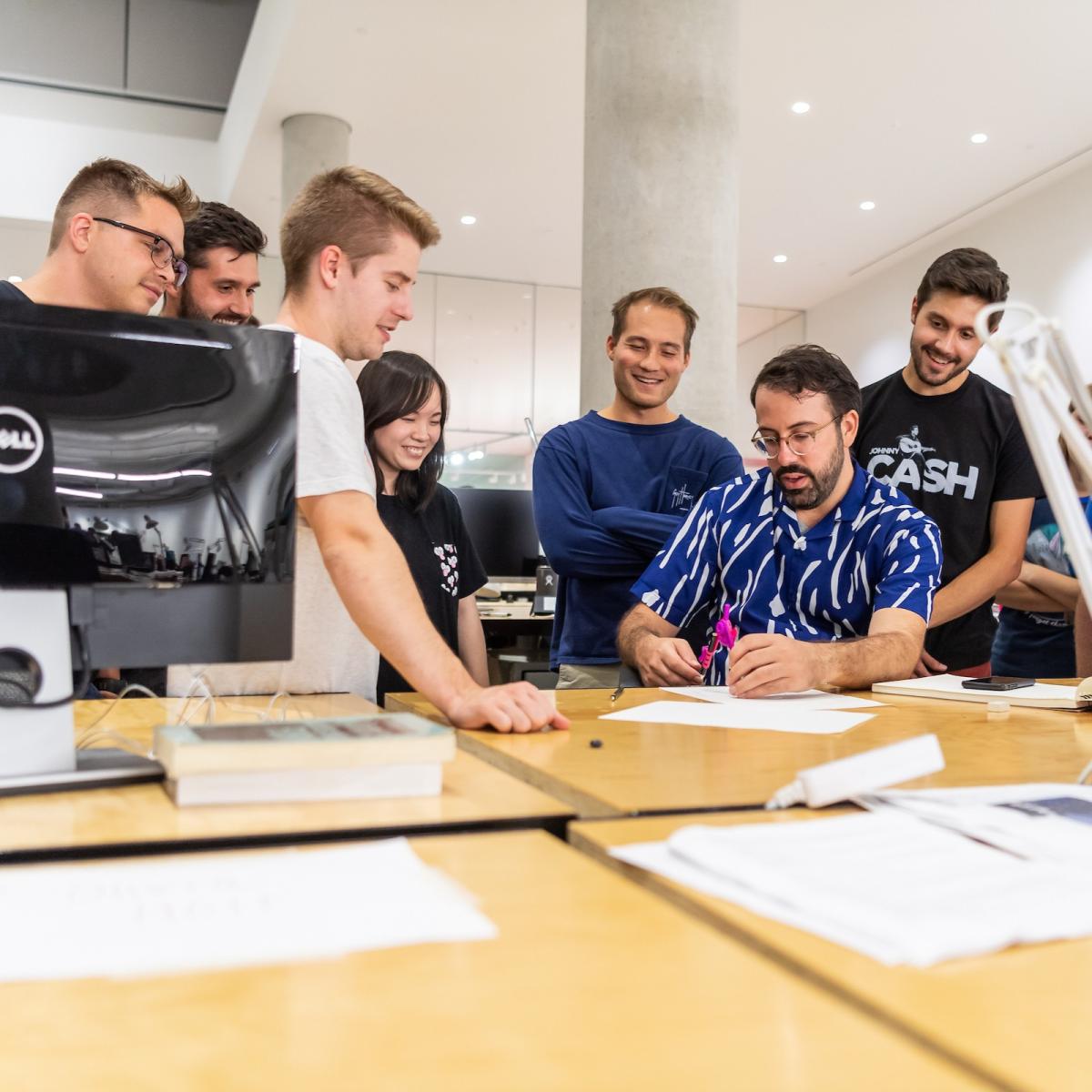 A group of students huddled around a desk where one student draws on a piece of paper