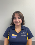 Meg W. with her short brown hair smiling at the camera, wearing her KSU blue and gold RecWell uniform shirt and RecWell nametage on her right side. Photo taken against a plain white wall. 