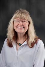 Photo of a woman (Amy Reynolds) in front of a gray background wearing a white button up, glasses, and a necklace. 