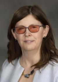 Photo of a woman (Alicia Crowe) with a white sweater on and amber glasses with a gray background. 
