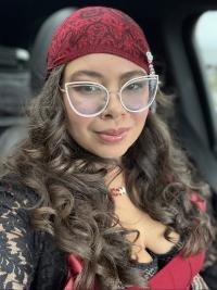 Portrait of Siera wearing a red hair wrap over long, wavy brown hair. She has a neutral expression and wears white cat-eye glasses 