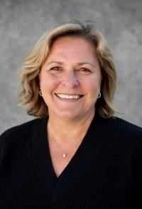 Photo of a blonde woman smiling in front of a gray background. Woman is wearing a black v-neck sweater shirt, pearl necklace, and earrings.