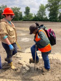 A woman is digging in the dirt with a shovel, wearing a hi-vis vest. Another woman stands in front of her, supervising, and wearing a hard hat. They are both in a barren landscape, there are trees far off in the background.