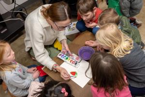 An art education student painting with watercolor while young students watch.
