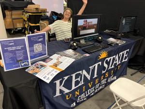 A Kent State University representative welcomes visitors at an Animation Game Design booth featuring a playable student game, program information and promotional materials.