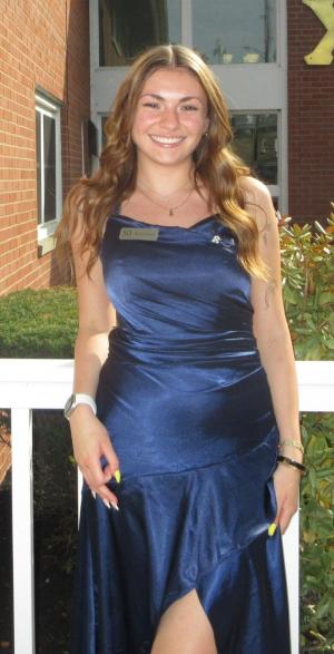 Person standing outdoors in front of a brick building, wearing a navy blue sleeveless dress with a thigh‑high slit and a name badge. The person has long wavy hair, a necklace, a wristwatch, and light-colored nail polish, with shrubs and windows visible in the background.