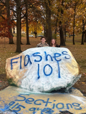 Two people standing behind a large painted rock in a park surrounded by autumn trees and fallen leaves. The rock is painted white with blue spray-painted text reading “Flashes 101,” with “Sections” partially visible on the ground in front of the rock. Trees, grass, and parked cars appear in the background.