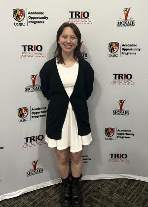 Person standing indoors in front of a step‑and‑repeat backdrop featuring logos for UMBC Academic Opportunity Programs, TRIO Ronald E. McNair Post‑Baccalaureate Achievement Program, and the McNair Scholars Program. The person is wearing a white dress with a black cardigan and black boots, standing on carpeted flooring against the branded background.