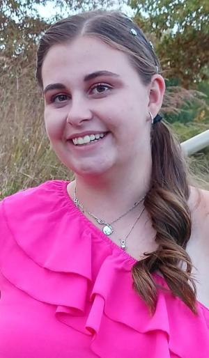 Melody outdoors wearing a bright pink one-shoulder ruffled top. Hair is pulled to one side with small hair clips, and she is wearing small hoop earrings and layered necklaces. The background shows tall grass and trees in daylight.