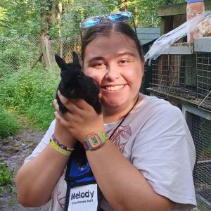 Melody outdoors holding a small black rabbit with both hands. She is facing the camera, wearing a light-colored T-shirt, a colorful digital watch, and sunglasses resting on their head. A name badge reads ‘Melody – Camp Wanake Staff.’ Wire animal enclosures and trees are visible in the background.