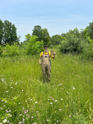 Brocato, outstanding in his field! He is literally standing in a field with tall grass and flowers, the grass is illuminated with sunlight and the sky is so clear and blue. He is wearing sunglasses and holding a field notebook, and smiling. He posed for the picture.