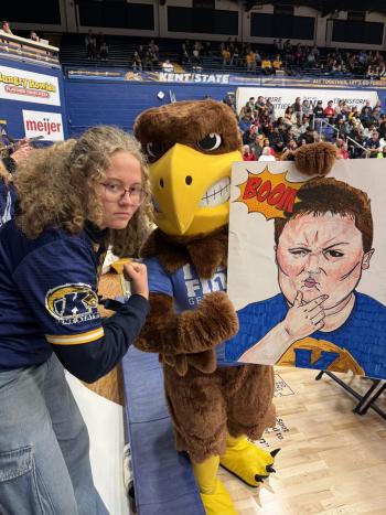 Bailey, wearing Kent State apparel, poses beside the Kent State eagle mascot on a basketball court, while the mascot holds a poster featuring a hand-drawn portrait with a comic-style “BOOM” graphic. Bleachers with spectators and Kent State signage are visible in the background.