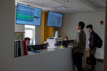 Two students across a counter looking at two large screens showing arrival and departure times for student flights.
