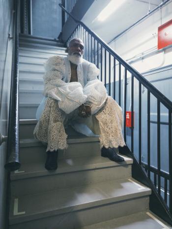 A man with a white beard sits on a set of stairs with his arms on his knees. He wears an all-white outfit of French lace and ruffled sleeves