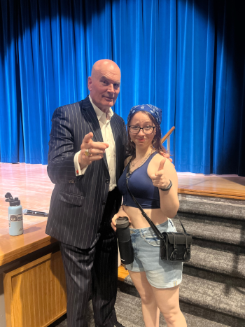Kay and Tim Misny, famous Cleveland personal injury lawyer, standing together on a stage with blue curtains in the background. One person is wearing a dark pinstriped suit and pointing toward the camera, while the other is wearing a sleeveless top, denim shorts, glasses, and a headscarf, holding a water bottle and giving a thumbs‑up. A set of steps, wooden stage flooring, and a water bottle placed on the stage are visible.