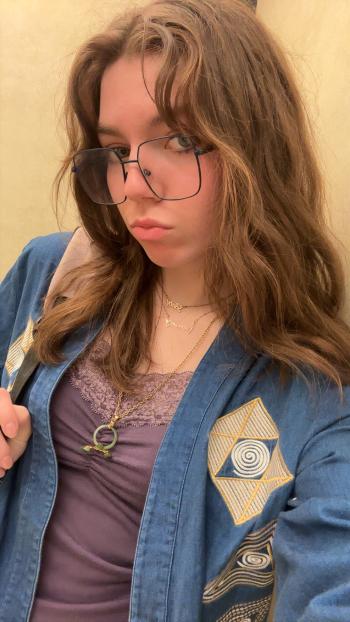 Close-up portrait of a person with long brown hair wearing oversized square glasses. The person is dressed in a blue jacket with geometric embroidered patches over a purple lace-trim top and layered necklaces, including one with a green circular pendant. The person is looking slightly to the side, with a neutral expression, against a softly lit indoor background.