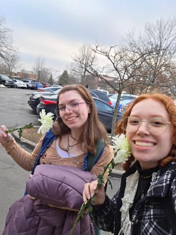Two people take a selfie outdoors in a parking lot lined with cars and leafless trees. Both are smiling toward the camera and each holds a white flower. One wears glasses and layered tops with a scarf and patterned outerwear, while the other wears glasses, a long-sleeve shirt with a vest, and carries a puffy jacket. The background shows overcast sky and a row of parked vehicles.