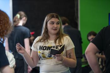 Sophia standing in front of green screen wearing a white tshirt, with a notebook in her hand