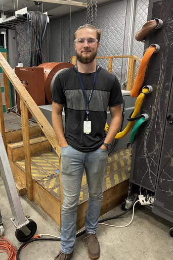 A young man wearing a dark t-shirt and jeans in a workroom.