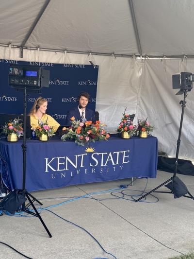 Two students sitting at a mobile broadcast desk with lights and cameras on them.