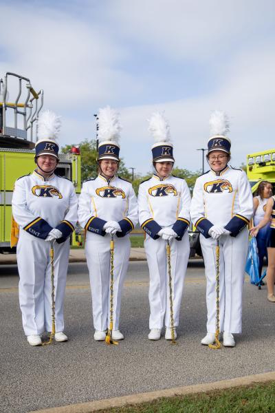 Four Kent State marching band members stand side by side on a street during a daytime event, wearing white uniforms with blue-and-gold accents and tall white plumes on their hats, each holding a ceremonial mace. A bright green fire truck and other parade participants are visible in the background under a partly cloudy sky.