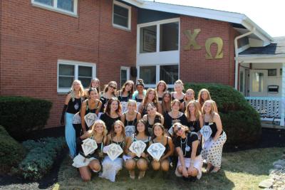 Large group posing outdoors in front of a red brick building with gold Greek letters “ΧΩ” mounted on the wall. The group is arranged in rows on grass and shrubs, wearing coordinated black and light-colored outfits, and several people are holding diamond-shaped signs with names written on them. A porch, windows, and landscaping are visible in the background under bright daylight.