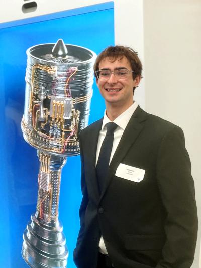 CAE student Ben Windt dressed professionally in a white button, black tie, and black suit coat and stadning next to a digital twin of a manufacturing piece at his internship.