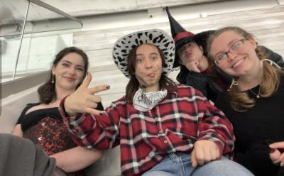 Four people sitting together indoors, leaning toward the camera and wearing themed or costume-style outfits. One person in the center wears a cowboy-style hat and plaid shirt and makes a playful hand gesture, while others wear dark clothing and hats. The group is seated against a light-colored wall with a glass railing visible, suggesting a public indoor venue.