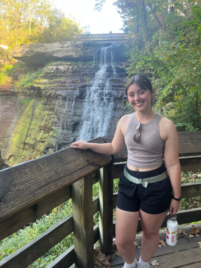 Olivia standing on a wooden overlook railing in front of a cascading waterfall surrounded by trees and greenery. The person is wearing a sleeveless top and shorts, holding a reusable water bottle, with a rocky cliff and a small bridge visible above the waterfall in the background.