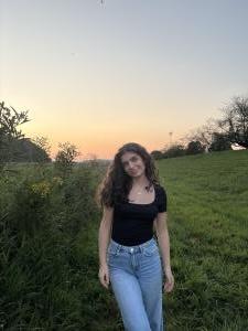 Zain Alkadi stands in a grassy field at sunset, wearing a black top and light blue jeans with trees and an open landscape behind her.