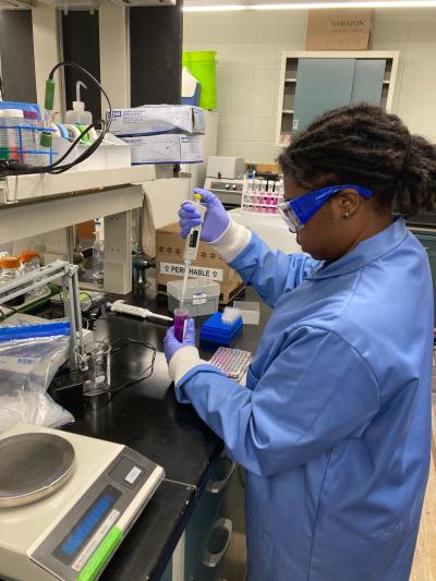 A woman is in a lab, wearing a lab coat and goggles. She is transferring a liquid from a pipette to a beaker.
