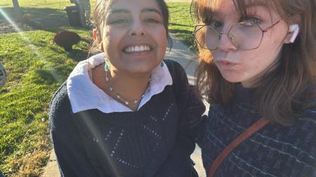 Two people pose closely together for an outdoor selfie in bright sunlight. One person wears a dark sweater layered over a white collared shirt, a beaded necklace, and dangling earrings, smiling toward the camera. The other person, wearing large glasses and an earbud, has shoulder-length hair and a crossbody strap visible, tilts their head and looks toward the camera. The background shows a grassy area with trees, a sidewalk, and soft sunlight creating lens flare.
