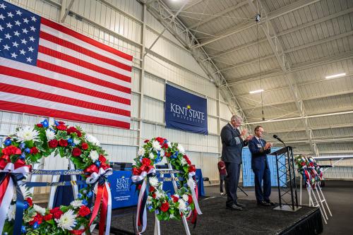 Two professionally dressed men at a podium with an American flag and a Kent State University flag hanging behind them. Red, white, and blue memorial wreaths are on the stage next to them.