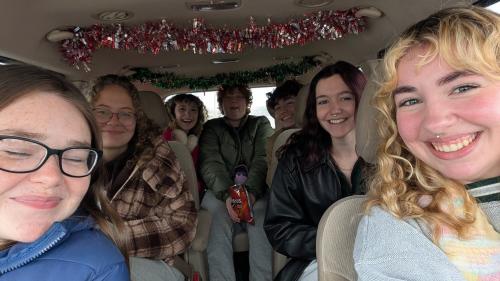 Bailey is on the right, part of a group of seven people inside a vehicle, facing the camera, with decorative red and green tinsel strung across the ceiling. The group fills the front and back rows of seats, wearing casual jackets and sweaters. One person in the center holds a bag of Doritos, and the interior of the vehicle is visible around them.