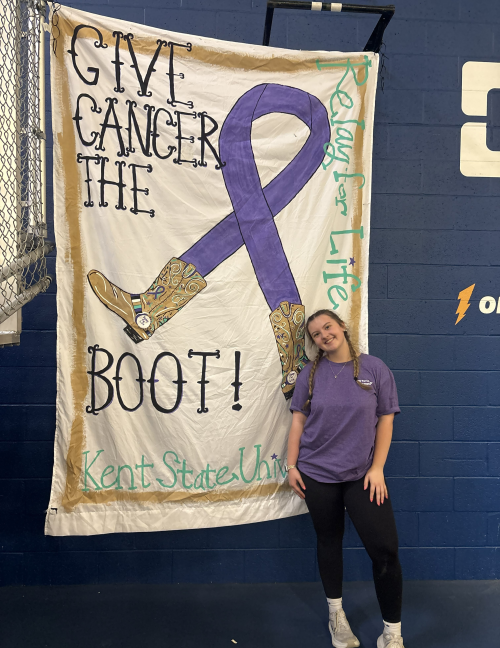 Isabella stands in front of a banner that reads "Give Cancer the Boot/Relay for Life."