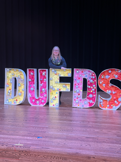 Kellyn stands on stage behind large letters spelling out DUFDS, the acronym for Dress Up For Downs Syndrome.