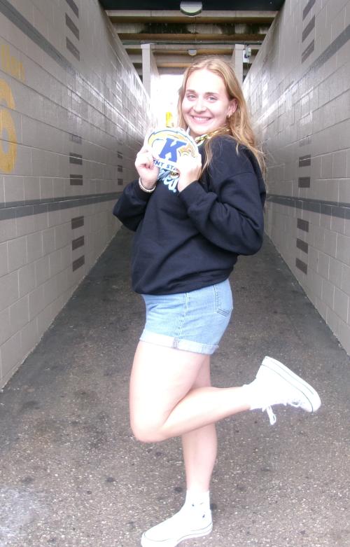 Norah poses in a hallway, holding a Kent State Athletics logo with a blue K in front of a lightning bolt.