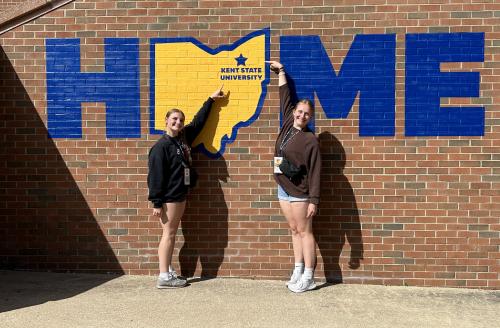 Norah and a friend stand in front of a painting on the wall of the Student Center. The painting is of the word HOME, with the O replaced by the shape of the state of Ohio.