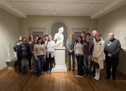 A group of about fifteen people stand in a museum gallery arranged around a white marble statue on a pedestal. The statue is centered in an arched alcove, with the group forming a semicircle on a wooden floor. Neutral-colored walls, display text panels, and gallery lighting are visible in the background.