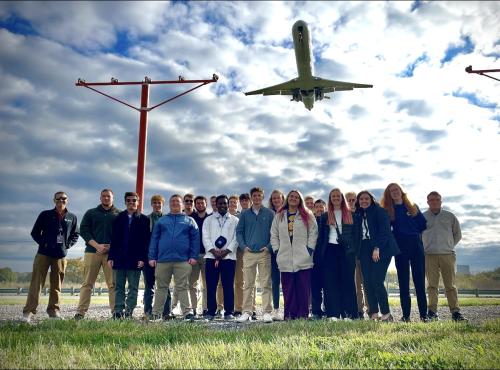 Zayden and his classmates pose for a group photo on an airfield while a plane flies overhead.