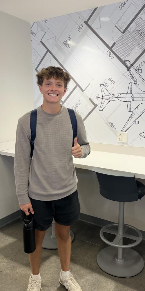 Zayden gives a thumbs up as he stands in front of a desk in the Aeronautics Building.